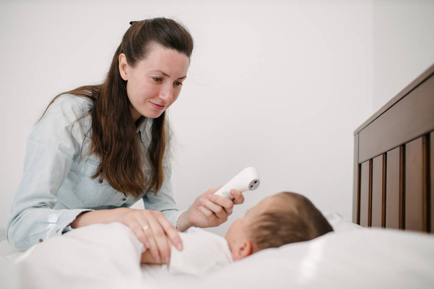 Η γυναίκα μετρά τη θερμοκρασία. Το παιδί είναι άρρωστο ξαπλωμένο στο κρεβάτι. - Φωτογραφία, εικόνα