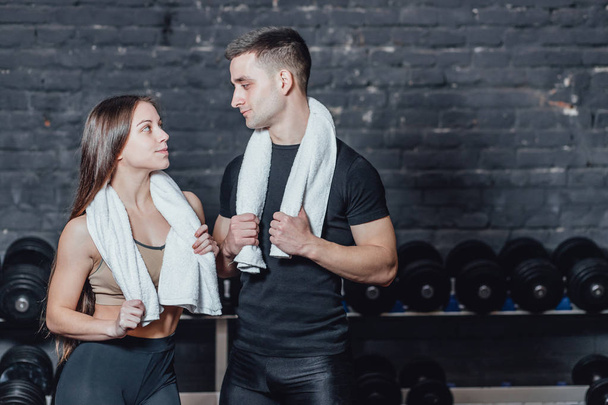 Young sporty couple guy and girl standing in gym. During this, the white scumbag is held on the neck. - Фото, изображение