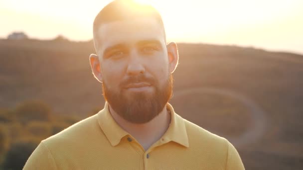 Close up, Portrait of a handsome Caucasian bearded young man looking straight to the camera on the background of a bright sunset over natural landscape. Joy of life, active lifestyle, blogger. - Materiaali, video