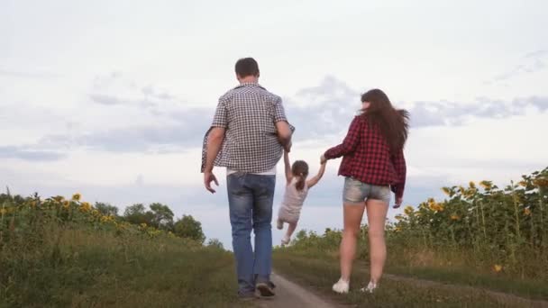 baby flies in the hands of father and mother. little daughter jumping holding hands mom and dad. Family with small child walks along road and laughs next to field of sunflowers. - Materiał filmowy, wideo
