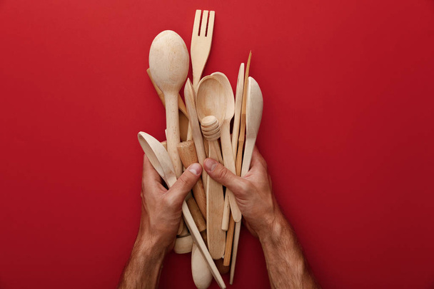 cropped view of man holding wooden spoons, fork and kitchenware on red background - Photo, Image