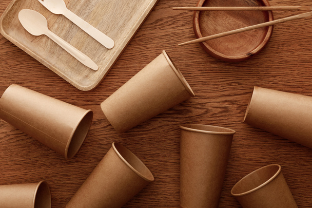 top view of wooden dishes, cutlery, chopsticks and paper cups on brown background - Photo, Image