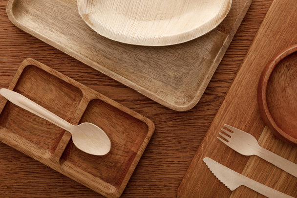 top view of wooden dish and cutting board with plates and cutlery on brown background - Foto, Imagen