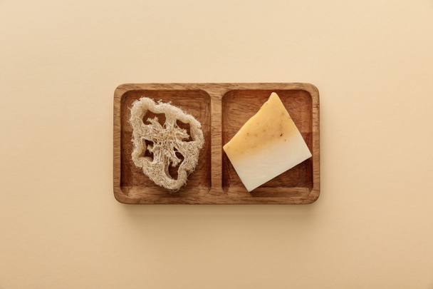 top view of brown wooden soapdish with loofah and piece of soap on beige background - Foto, Imagen