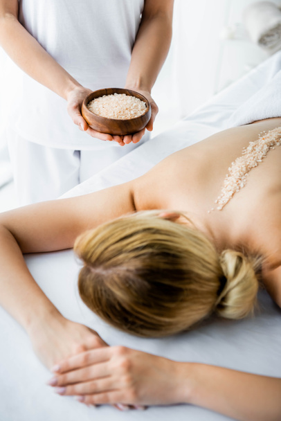 przycięte widok z masażysta posiadania miski z solą morską i kobietą leżącą na mat masażu w spa  - Zdjęcie, obraz