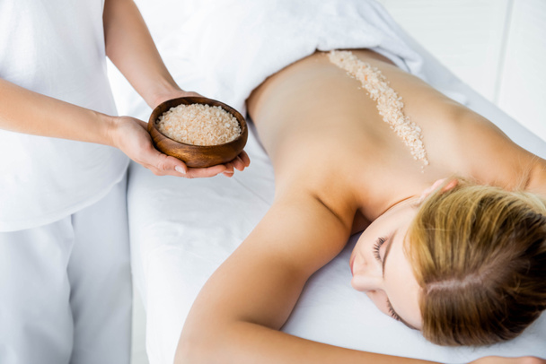 vista recortada del masajista sosteniendo tazón con sal marina y mujer acostada en la alfombra de masaje en el spa
  - Foto, Imagen