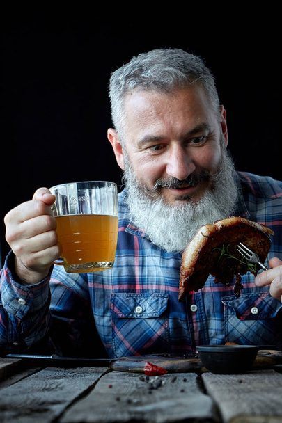 Brutální šedovlasý dospělý muž blázen do hořčičného steaku a piva, koncept dovolené, festivalu, Oktoberfestu nebo den svatého Patrika - Fotografie, Obrázek
