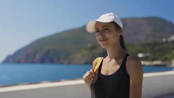 outside portrait of Beautiful stylish sexy sporty woman in black swimsuit and white cap walking along bay at seaside, against a blue sea at sunny day then smiling at camera. - Séquence, vidéo