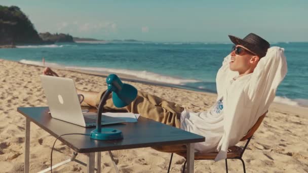Mladý muž sedí na pláži s laptopem, odpočívá a položil nohy na stůl - Záběry, video