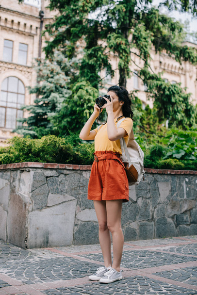  κορίτσι κρατώντας ψηφιακή φωτογραφική μηχανή, ενώ τη λήψη φωτογραφιών κοντά στο κτίριο και τα δέντρα  - Φωτογραφία, εικόνα