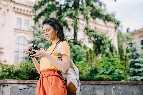 κορίτσι κρατώντας ψηφιακή φωτογραφική μηχανή κοντά στο κτίριο και τα δέντρα  - Φωτογραφία, εικόνα