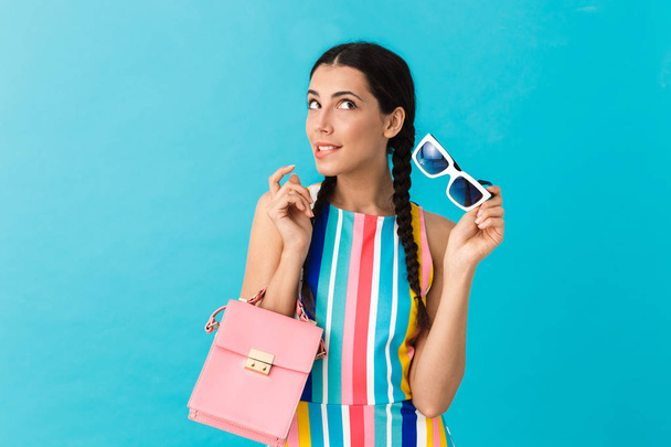 Image of brunette puzzled woman with pigtails biting her lip while holding pink bag and sunglasses - Photo, Image