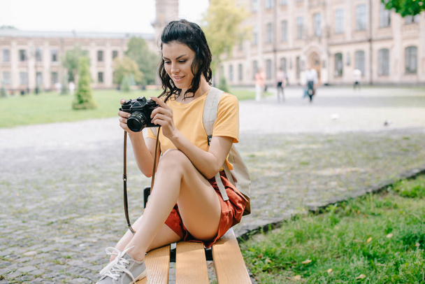 jovem atraente olhando para a câmera digital enquanto sentado no banco perto da universidade
  - Foto, Imagem