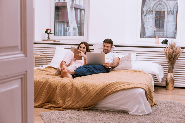 Man and his girlfriend in the morning in bed , cozy design , reading book and looking on the laptop - Photo, Image