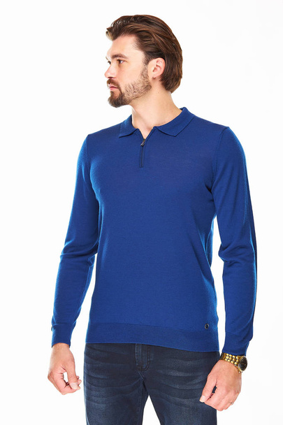 man with beard and dark hair stands on a white background in a blue sweater with a zip and dark jeans, he has a gold watch on his arm and looks away - Foto, Bild