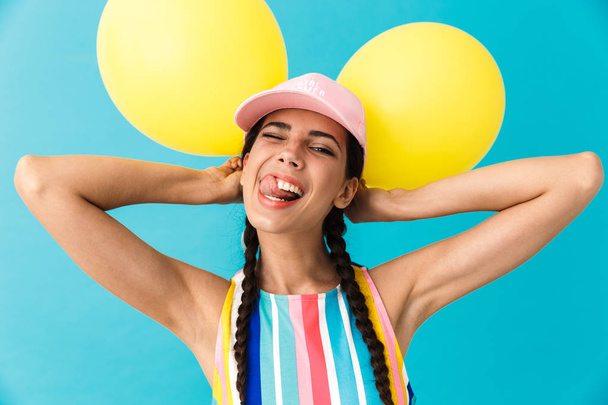 Image of young joyful woman wearing cap winking while holding two air balloons - Фото, изображение