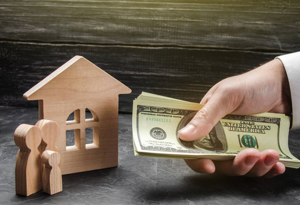 The hand of a businessman stretches a wad of money to the family figures near the wooden house. Providing a mortgage loan for buying a home and improving home conditions. Buying a house - Photo, image