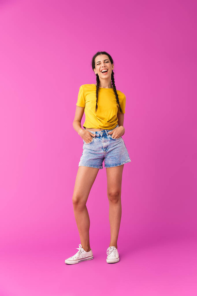 Full length photo of young joyful woman wearing denim shorts winking with her hands in pockets - Photo, image