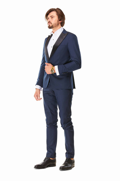 man with beard and dark hair stands on a white background in dark blue tuxedo, white shirt and black leather shoes, he has a gold watch on his arm and he looks awa - Photo, Image