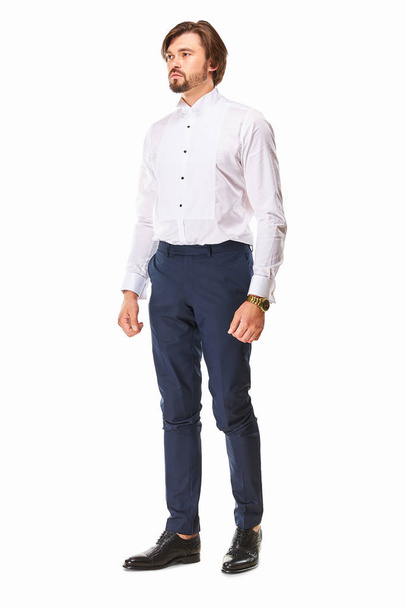 man with beard and dark hair stands on a white background in white shirt for  tuxedo, dark blue trousers and black leather shoes, he has a gold watch on his arm and he looks awa - Foto, imagen