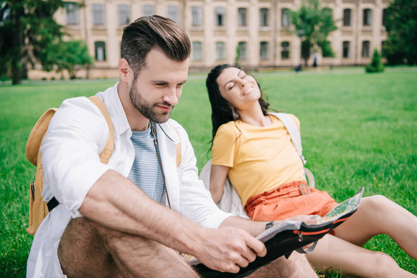 Selektiver Fokus eines glücklichen Mannes, der Landkarte in der Nähe einer Frau hält, die auf Gras sitzt  - Foto, Bild