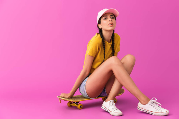 Image of seductive caucasian woman wearing casual clothes looking at camera and sitting on skateboard - 写真・画像