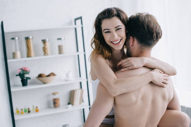 back view of boyfriend holding smiling and attractive girlfriend in apartment  - Fotografie, Obrázek