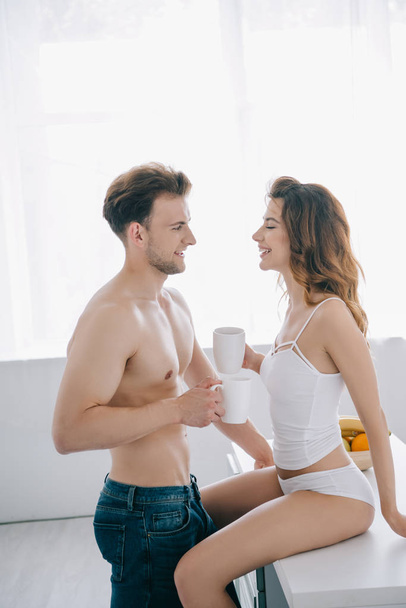 attractive girlfriend and handsome boyfriend smiling and holding cups with tea in apartment  - Foto, Bild