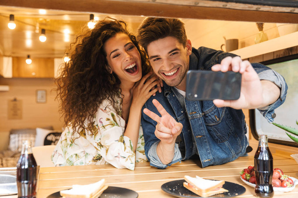 Retrato de pareja feliz gesto signo de paz mientras toma foto selfie en el teléfono inteligente en la acogedora cocina en el remolque en el interior
 - Foto, imagen