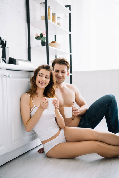 attractive girlfriend and handsome boyfriend smiling and holding cups in apartment  - Φωτογραφία, εικόνα