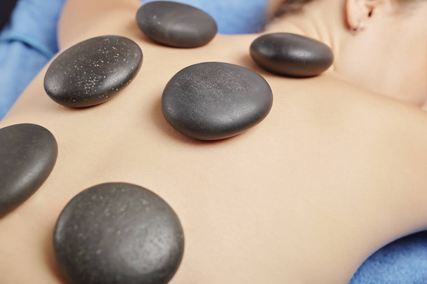 Smooth heated basalt stones on back of young female spa salon client, selective focus - Valokuva, kuva