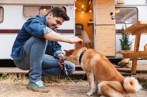 Portrait of unshaven handsome man wearing denim clothes stroking red dog while squatting near house on wheels outdoors - Фото, зображення