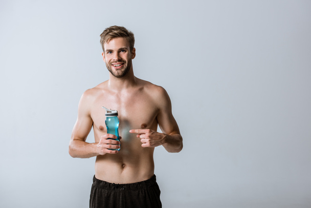 front view of smiling shirtless bearded sportsman pointing with finger at sport bottle isolated on grey - Foto, Imagen