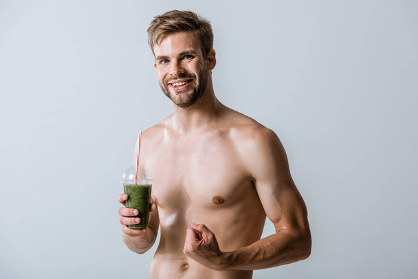 smiling shirtless sportsman holding smoothie and clenching fist isolated on grey - Photo, Image