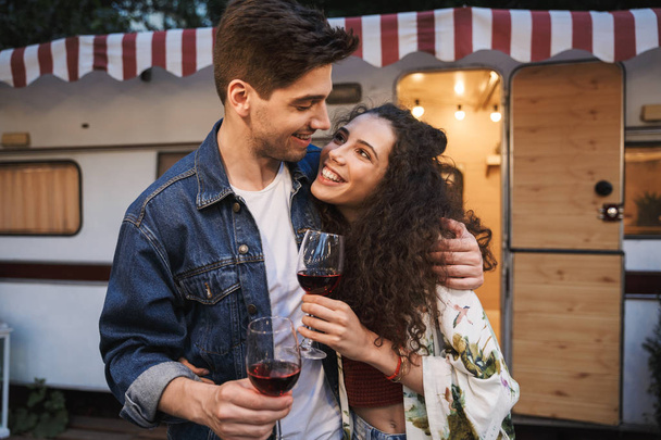 Portrait of satisfied couple drinking red wine and hugging together while standing near trailer outdoors - Foto, Imagem