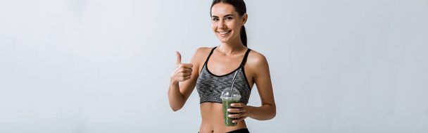 panoramic shot of smiling sportswoman holding smoothie and showing thumb up isolated on grey - Fotó, kép