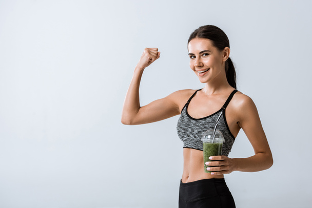 beautiful smiling sportswoman holding smoothie and showing biceps isolated on grey - Foto, Imagem