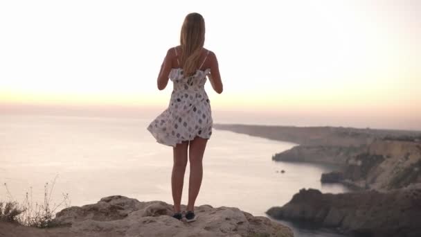 Footage from the backside of young, blonde girl standing on the cliff and admiring the view from the cliff. Carefree, cheerful girl in sunset light with outstretched hands, wind blowing her dress and - Footage, Video