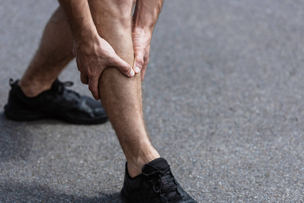 partial view of sportsman in black sneakers with leg pain on street - Foto, Imagem