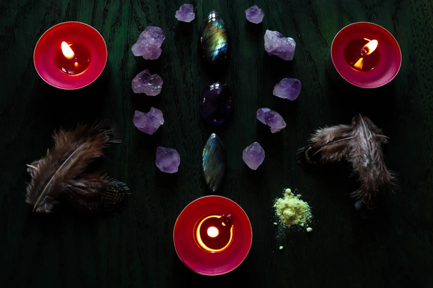 Magical layout of stones (labradorite, amethyst), feathers, sulfur and red candles on a green wooden background. - Фото, изображение