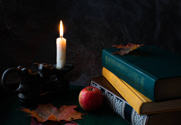 Autumn still life on a dark background with books, a vintage candlestick with a burning candle, leaves and a red apple. - Photo, image