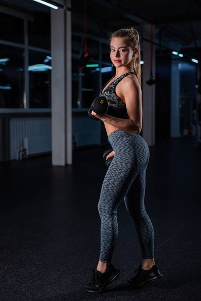 Beautiful muscular young woman doing exercises with dumbbells in the gym. The girl is working out the muscles of the arms with a dumbbell. Female bodybuilder trains biceps and triceps - Photo, image