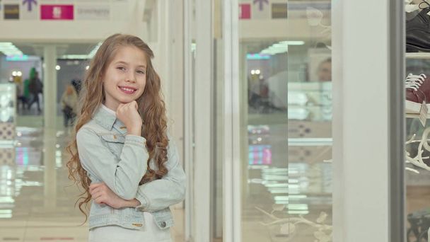 Милая маленькая девочка улыбается в камеру, осматривает витрину модного магазина
 - Фото, изображение