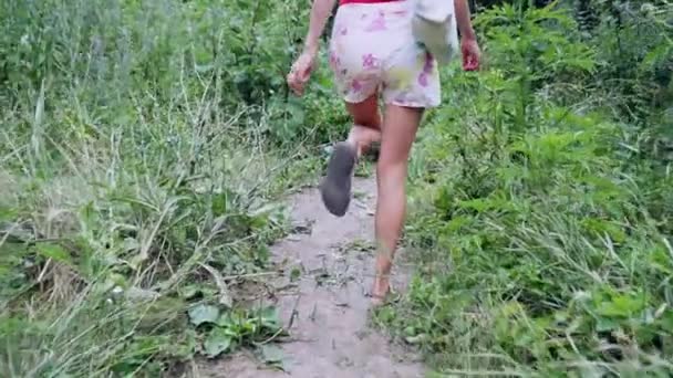 ブラウスとデニムショートパンツの裏側の女の子は、緑の森を通ってパスに沿って歩きます。スローモーション. - 映像、動画