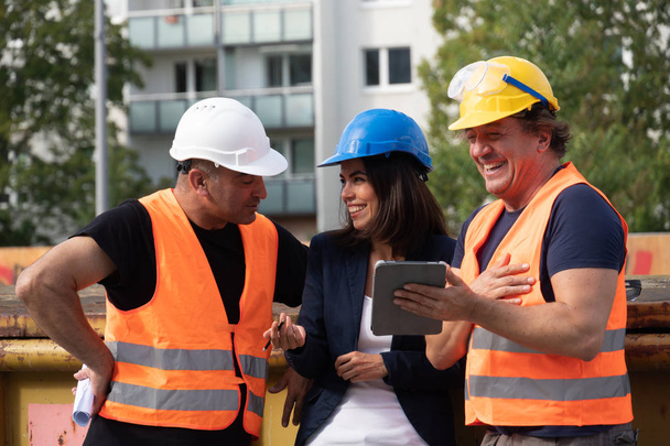 Foreman constructor con chaleco de seguridad, casco y gafas riendo con un hombre y una mujer colegas en el sitio de construcción
 - Foto, Imagen