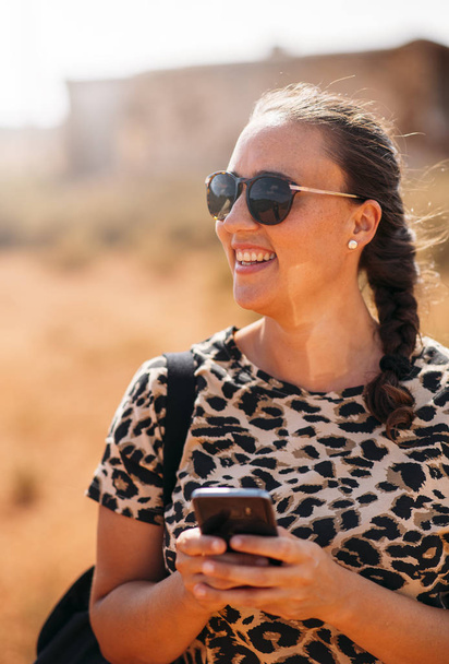 Close up of a beautiful young woman using her smart phone outside. Cheerful woman smiling with sunglasses, braid and animal print shirt. - Zdjęcie, obraz