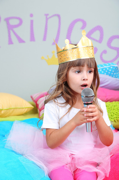 Little girl sitting on bed with microphone in room on grey wall background - 写真・画像