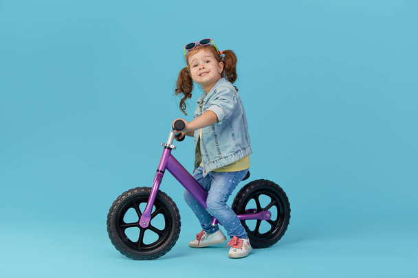 Little girl learns to keep balance while riding a bicycle. Stylish child in sunglasses  isolated on blue background - Fotó, kép