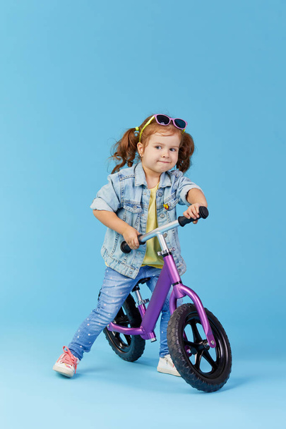 Little girl learns to keep balance while riding a bicycle. Stylish child in sunglasses  isolated on blue background - 写真・画像