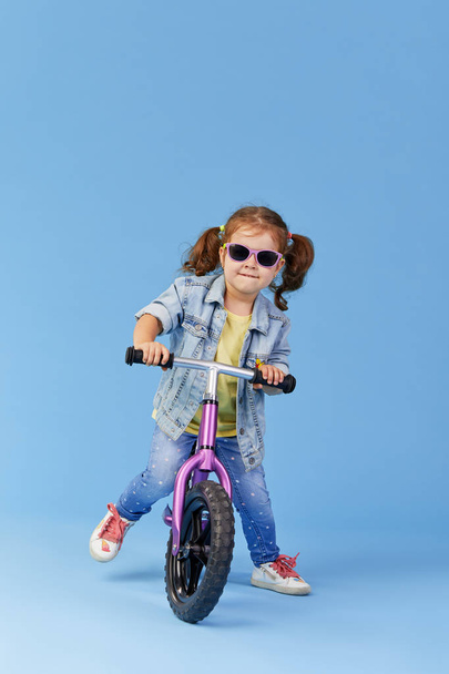 Little girl learns to keep balance while riding a bicycle. Stylish child in sunglasses  isolated on blue background - Foto, Imagem